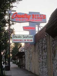 A stone and brick building with a white sign that reads "Country Bill's Restaurant" and "Cocktails" in red neon lighting, then "Steaks" and "Seafood" in black text, followed by "Parking in Rear" in red text