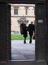 The tranquillity of an Oxbridge college glimpsed through an open wicket gate in the outside oak door. Paving stones lead to a grass quadrangle in front of an old two-storey building in yellow-pink stone, with sash windows on the upper floor above a passage entrance decorated with rococo carving and painted crest, leading to another grassed quadrangle. A male and female student, similarly dressed in short black coats, are walking in step away from the gate and into the depths of the college carrying their bags and holding hands.