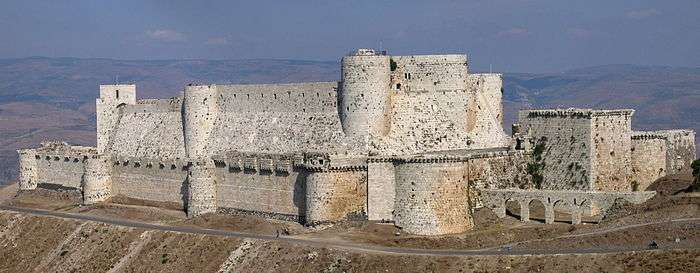 A stone castle with two high curtain walls, one within the other. They are crenelated and studded with projecting towers, both rectangular and rounded. The castle is on a promontory high above the surrounding landscape