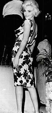 Monroe wearing a form-fitting white dress with flowers and an open back. She is standing and smiling over her shoulder at the camera.