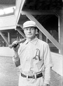 A man wearing an old-style white baseball uniform with a large script "A" over the left breast and holding a baseball bat over his right shoulder