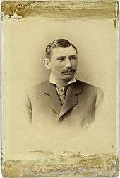 A sepia-toned image of a mustachioed man in a dark Victorian suit