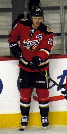 A hockey player in full uniform. The Jersey and legs are red with black, white and yellow trim, while the pants and helmet are black.  The logo says "Calgary" in script and includes a small stylized "C".