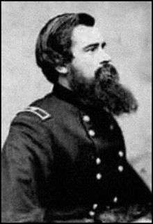 Profile of a white man with a long beard and thick hair wearing a double-breasted military jacket with a rectangular patch on the shoulder.