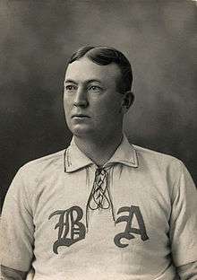 A black-and-white photograph of a man from the chest up looking to his right, wearing a baseball uniform with the letters "B" and "A".
