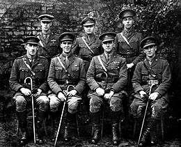 Formal portrait of seven men in military uniforms with peaked caps, four seated and holding canes, and three standing behind them