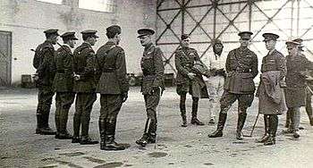A group of military personnel in an aircraft hangar, four of whom are in a row facing another man, while the remainder stand informally, one of them wearing a flying suit