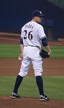 A man wearing navy blue cap and a white baseball uniform with his last name, "Parra", and the number 26 written on the back in navy blue letters stands on the pitcher's mound