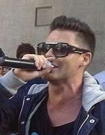 Man with spiked-up black hair and wearing black sunglasses and jacket singing into a hand-held microphone