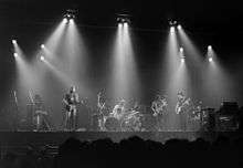 A monochrome image of Pink Floyd performing on a concert stage. Each band member is illuminated from above by bright spotlights