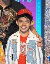 A smiling, young Indian boy wearing a red T-shirt, blue and white jacket, and red hat.