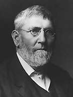 A middle-aged man with a beard and old-fashioned wire-framed glasses. His head and shoulders are visible, and he is turned three-quarters of the way towards the right. He is wearing a dark suit and waistcoat with a white shirt.