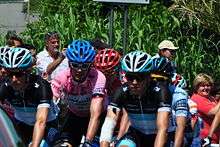 A group of cyclists with sad expressions on their faces riding together. Prominent is one wearing a pink jersey. Spectators watch from the roadside.