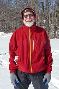 A man with a white beard, mustache and sunglasses, wearing a hooded red jacket with yellow zipper, and blue jeans, photographed in winter with snow and bare trees behind him