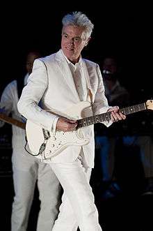 David Byrne looking down at the crowd and smiling while playing guitar