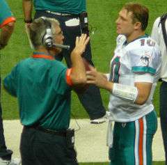 Candid photograph of Lee wearing a green polo shirt and headset standing on a football sideline and gesturing with his right hand in a conversation with Chad Pennington who is wearing a Miami Dolphins uniform