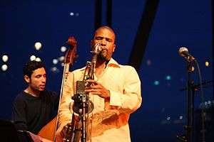 A black man in the front wearing a light orange shirt and playing the saxophone with his eyes closed and in the back a white man with his eyes closed playing the cello.