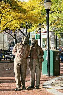 A sculpture resembling an elderly man and woman walking down the street, with trees and buildings in the background.