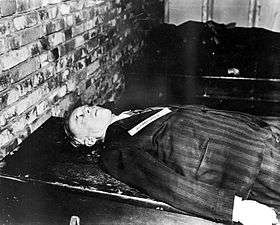 A corpse dressed in a black suit lies facing up on a table next to a brick wall. Only the upper torso is visible.