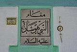 A closeup of a plaque with large Arabic script. There is more Arabic script inside a small gold-framed picture to the left of the plaque and a golden octagonal amulet hanging to the right of it.