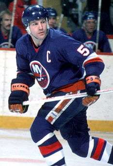 Denis Potvin wearing a helmet and hockey jersey, holding a hockey stick and skating