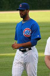 A dark skinned man in white pinstriped pants, a blue baseball jersey, and a blue baseball cap. The jersey and cap each have a red "C" on them.