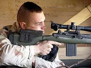color photo of a Marine peering through the optics of a large rifle