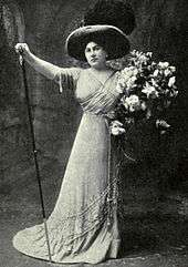  Woman standing in a dramatic pose, right arm raised, left arm holding a large bouquet. She is wearing a long formal gown and a wide-brimmed hat.