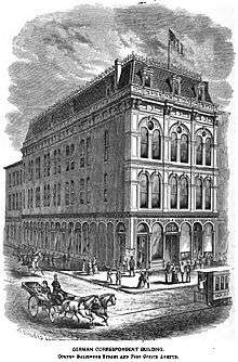 A lithograph of a four story building on the corner of two streets. It has tall, narrow arched windows in pairs with considerable decoration and a mansard roof. The stone street is busy with people, including a trolly partly out of frame and a two horse carriage.