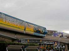 A monorail stamping a painted Light Cycle which leaves an orange trail behind. The second cart also has the Tron Legacy title.