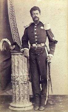 A photograph showing a mustachioed man in military uniform standing next to a truncated column upon which rests a military-style plumed hat