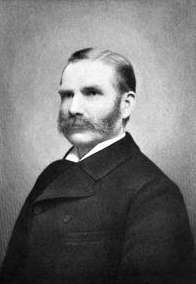 Formal half-length portrait of a middle-aged man with long sideburns, a full mustache, and neatly combed hair parted in the middle. He is wearing a dark suit and a white collar.