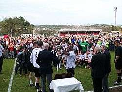 Two men in white football shirts and black shorts receive a silver trophy from an elderly man in a dark blazer.  More players look on, as do several more men in blazers and a crowd of spectators of all ages.  Several photographers are taking pictures of the presentation.