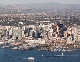 An overhead view of a San Diego's downtown with tall skyscrapers present throughout the area of different colors and sizes. A bay can be seen at the bottom of the image with several boats in the water. In the background are other neighborhoods near the downtown area as well as mountains in the distance.