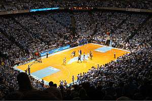 The inside of a basketball dome with two teams on a court with a large logo in the shape of the state of North Carolina in the middle.