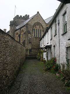 Stone building with arched windows and square tower seen at the end of a narrow lane with white painted houses on the right and a wall on the left.