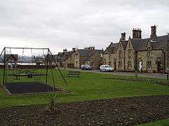 A village green with a swing set in the middle and a row of houses in the background
