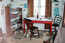 In the centre of a cluttered, whitewashed room stands a red table with two chairs, behind is a double window with simple brown curtains. Writing paraphernalia is strewn across the room