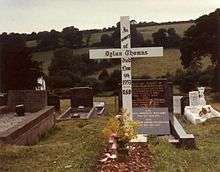 A simple white cross engraved with a memorial message to Thomas stands in a grave yard surrounded by hills