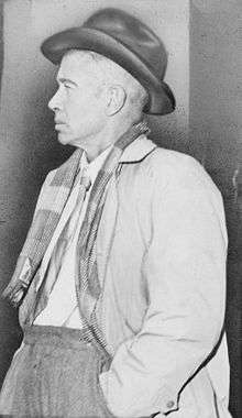A black and white photograph of a man in late middle-age wearing a dark cowboy hat, white shirt, tie, gray jacket, scarf and gray trousers. He faces left and his left hand is in his trouser pocket.