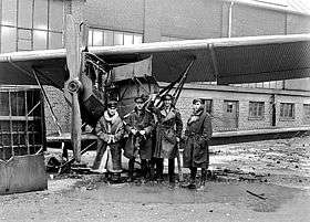 Four men in military uniforms with overcoats, standing next to a biplane parked in front of a building