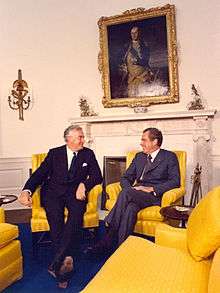 Whitlam sits in a chair, smiling, with Richard Nixon (whose appearance is well known) in another, who is also smiling.
