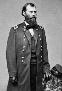 A white man with a full, bushy beard standing with his left hand resting on a table beside him; a slouch cap is on the table. He is wearing dark gloves, a long unbuttoned military jacket over a vest, and a bow tie.
