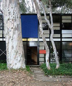 Photograph of the Eames House, a boxy building with a face of rectangles of various colors and clear glass, shaded by tall eucalyptus trees.