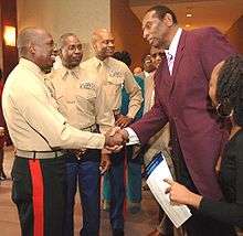 A tall African American man wearing purple suit is shaking hand with another African American man wearing a uniform, while three other men is watching on the background.
