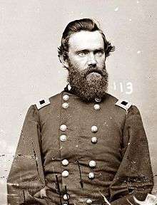 Head and torso of a white man with thick dark hair and a full beard, wearing a double-breasted military jacket with a rectangular patch atop each shoulder.