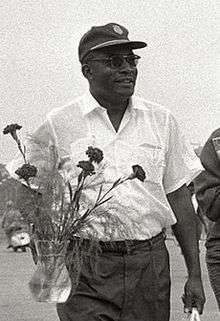 A black and white photo of a smiling man wearing a light-colored shirt and dark pants and hat carrying a vase of flowers.