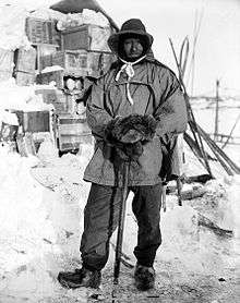 Full length picture of a man wearing heavy, Arctic clothing. He is standing outside in front of a snow-covered stack of wooden crates