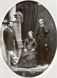 Three people: from left to right, a moustachioed gentleman of Mediterranean appearance, an older woman sitting on a chair, and a Catholic priest.