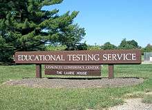 Large wooden sign with "Educational Testing Service" in white letters, in the middle of a field overlooking several trees and a blue sky.